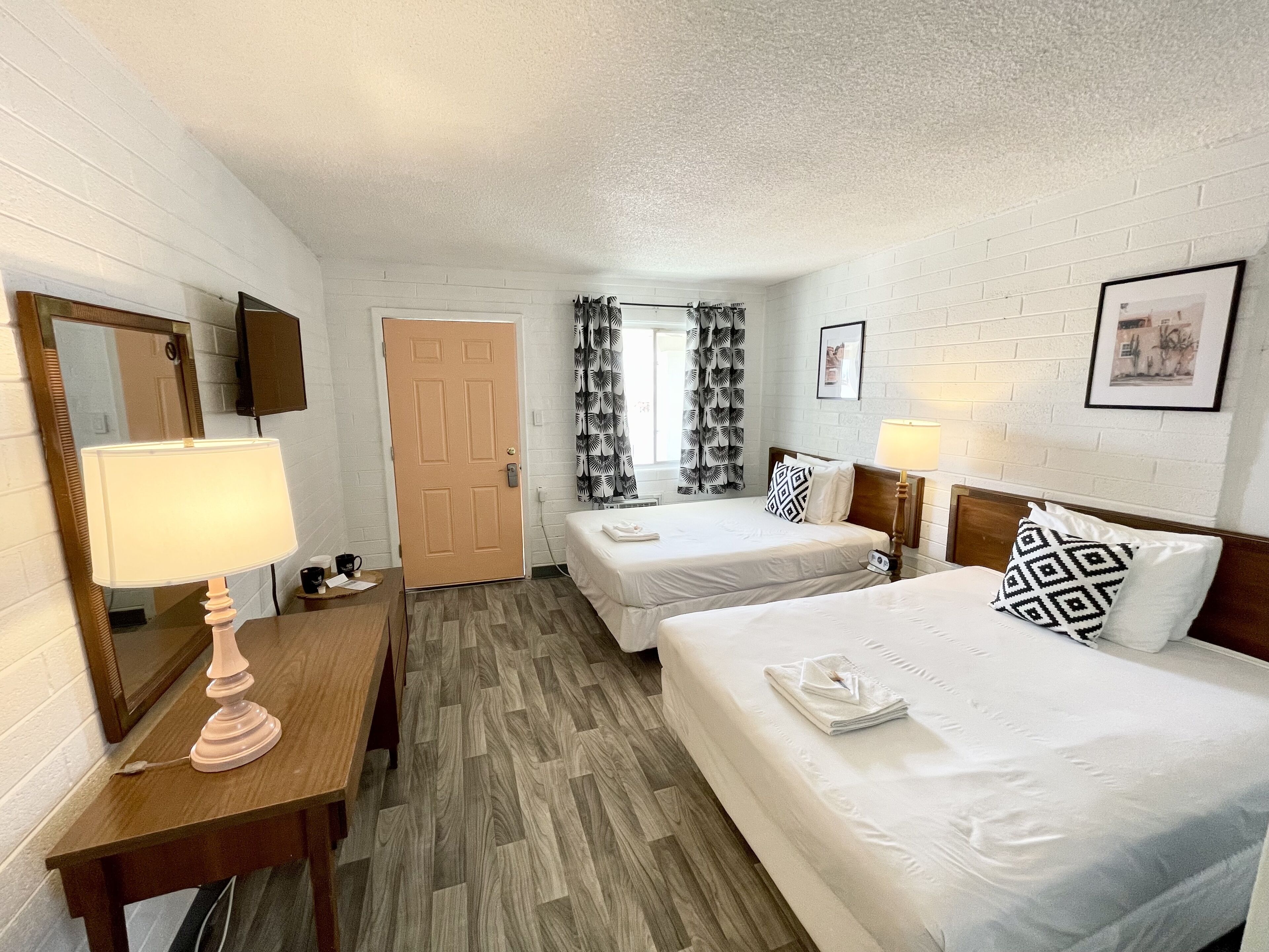 Habitación de hotel con dos camas, un escritorio de madera con lámpara, un espejo y una puerta.