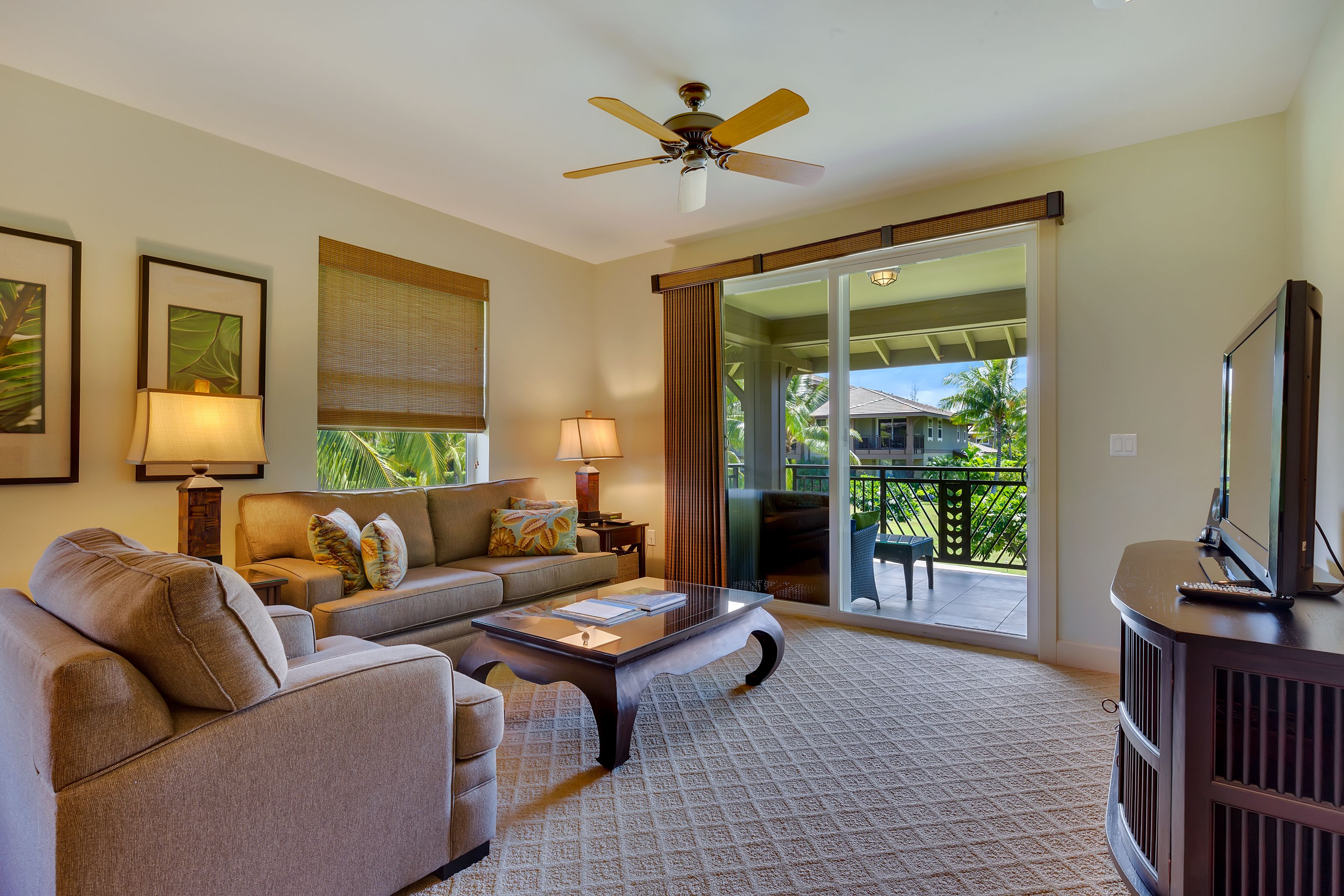 A living room with a sofa, coffee table, and a television on a stand. There is a ceiling fan and a view of the outdoors through a glass door.