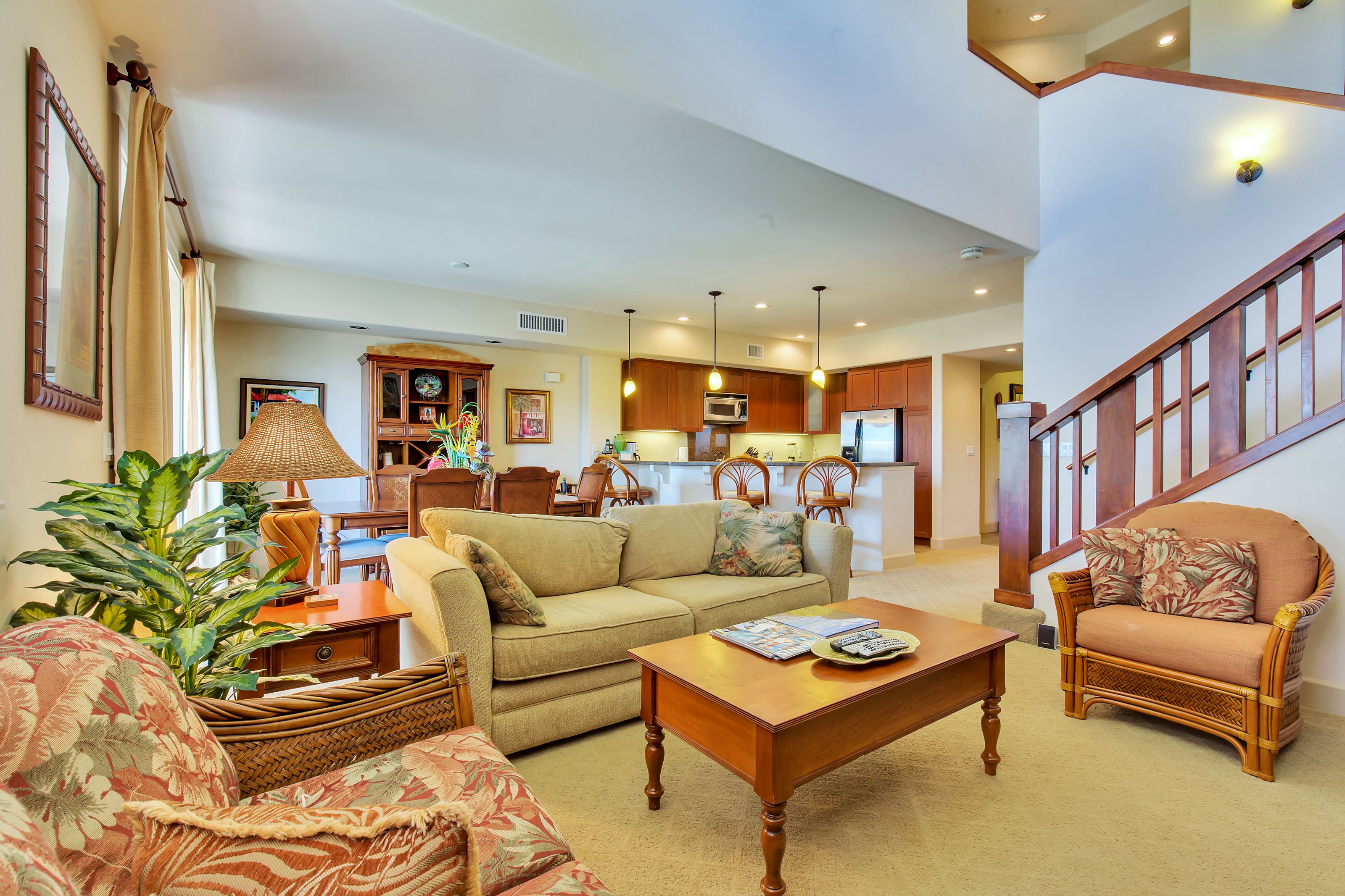 A spacious living room with a sofa, coffee table, and armchairs, a kitchen area with wooden cabinets and a microwave, and a staircase with wooden railings.
