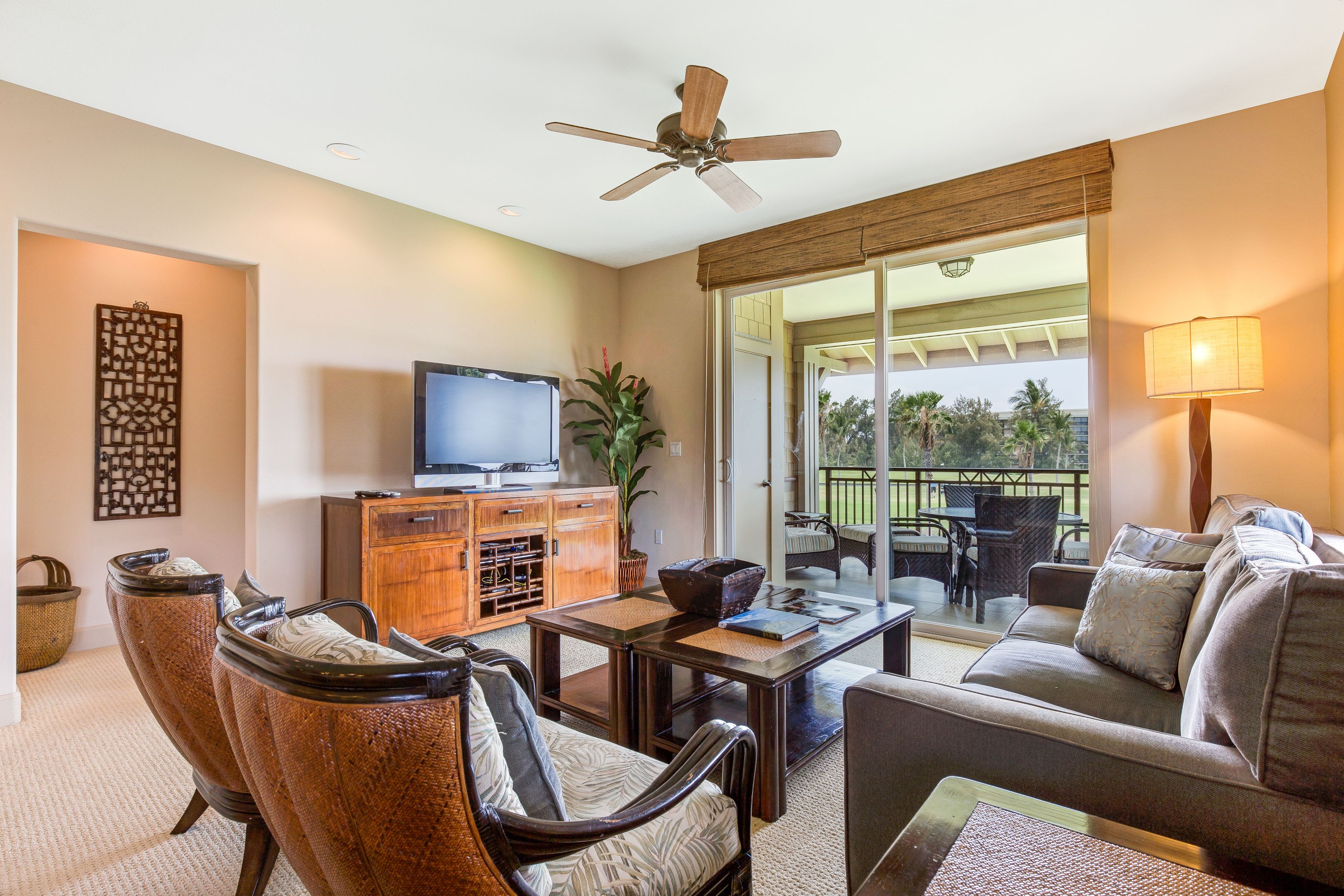 A living room with a television, wooden cabinet, wicker chairs, a sofa, a ceiling fan, and a view of the outdoors.
