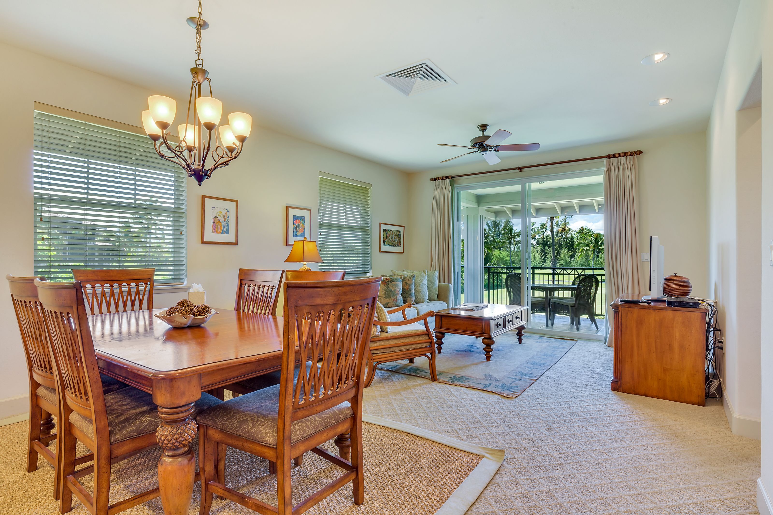 A dining area with a wooden table, chairs, and a chandelier. There is a sofa and a desk with a lamp.
