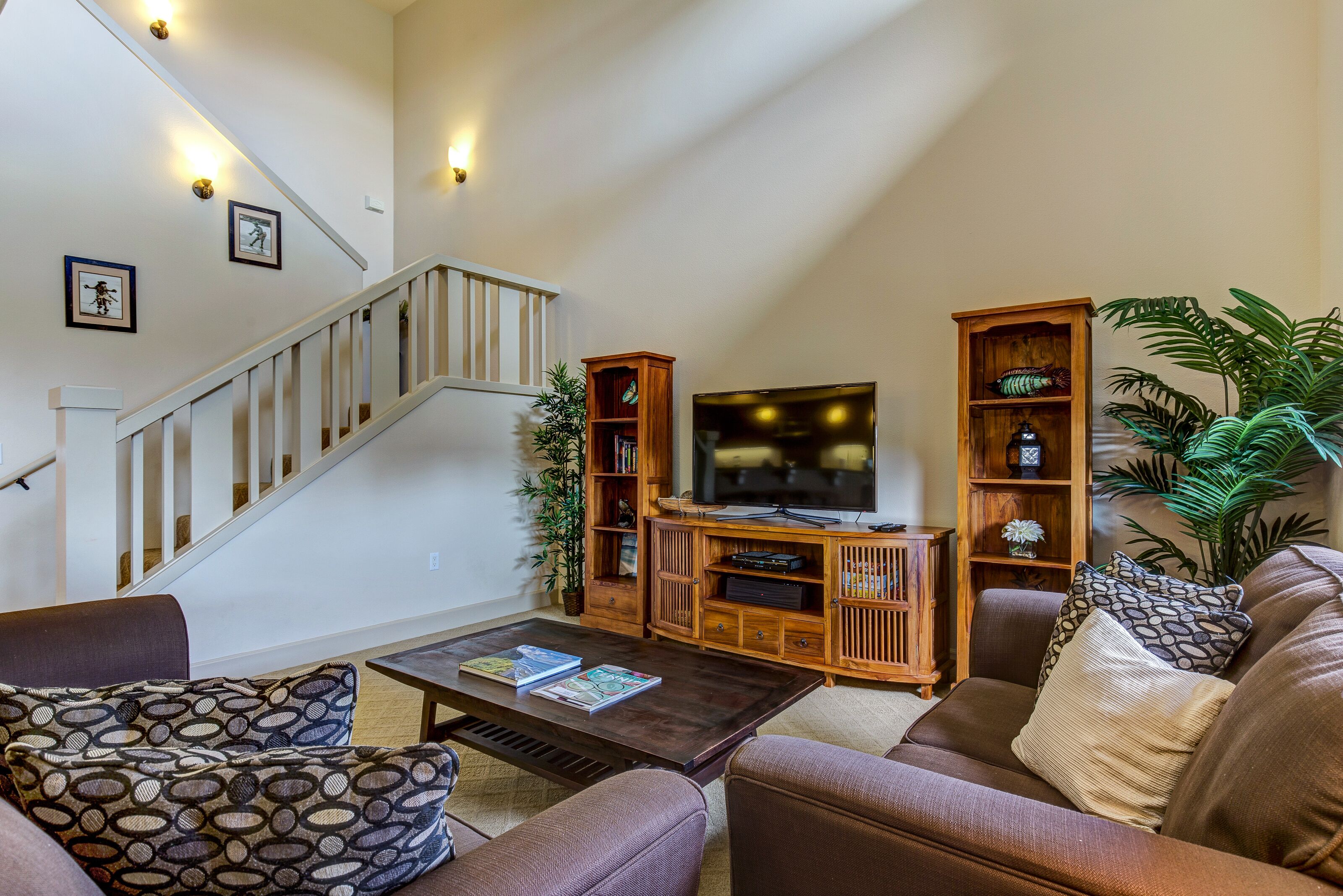A living room with a staircase, a flat-screen TV, wooden shelving, and a large potted plant.