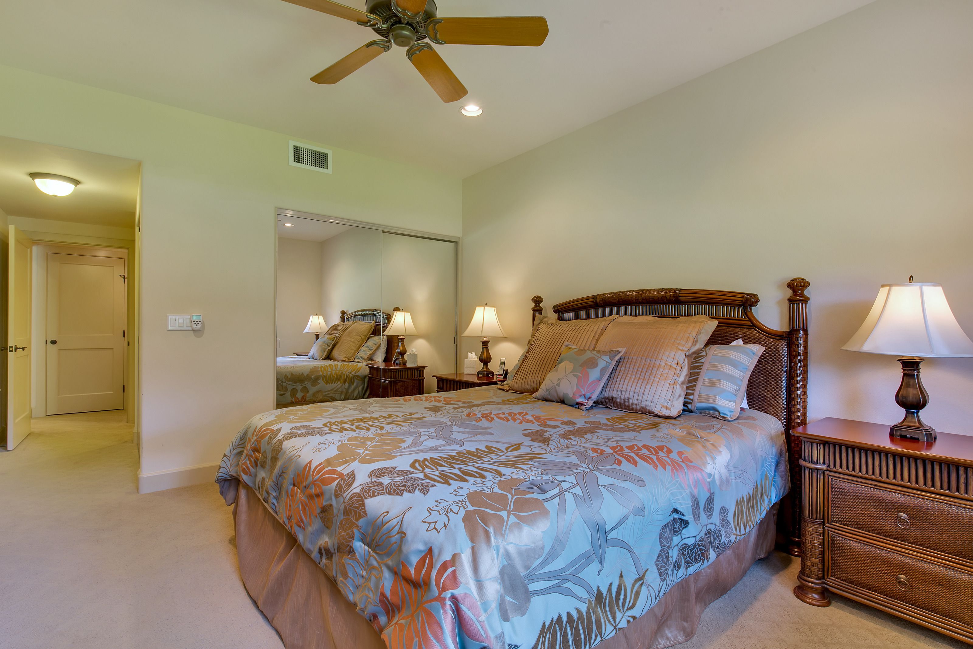 A bedroom with a large bed, two bedside tables with lamps, a ceiling fan, and a door leading to another room.