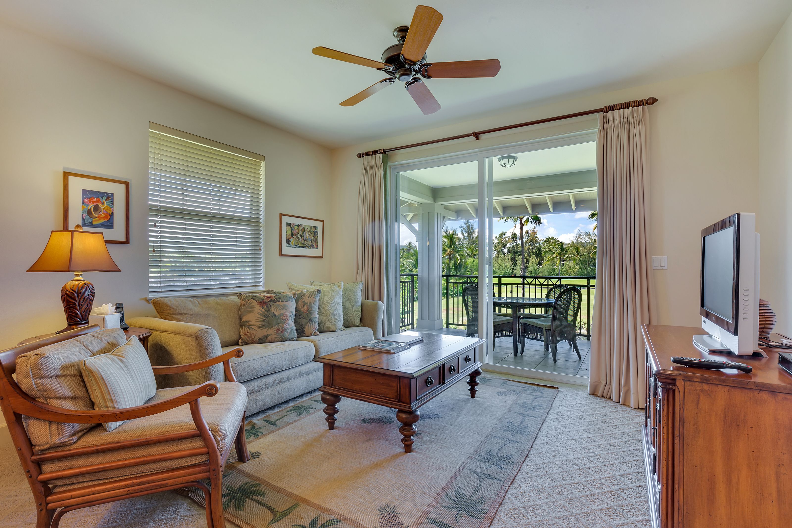 A living room with a sofa, coffee table, and a television on a wooden cabinet. There is a ceiling fan, a window with blinds, and a view of the outdoors.