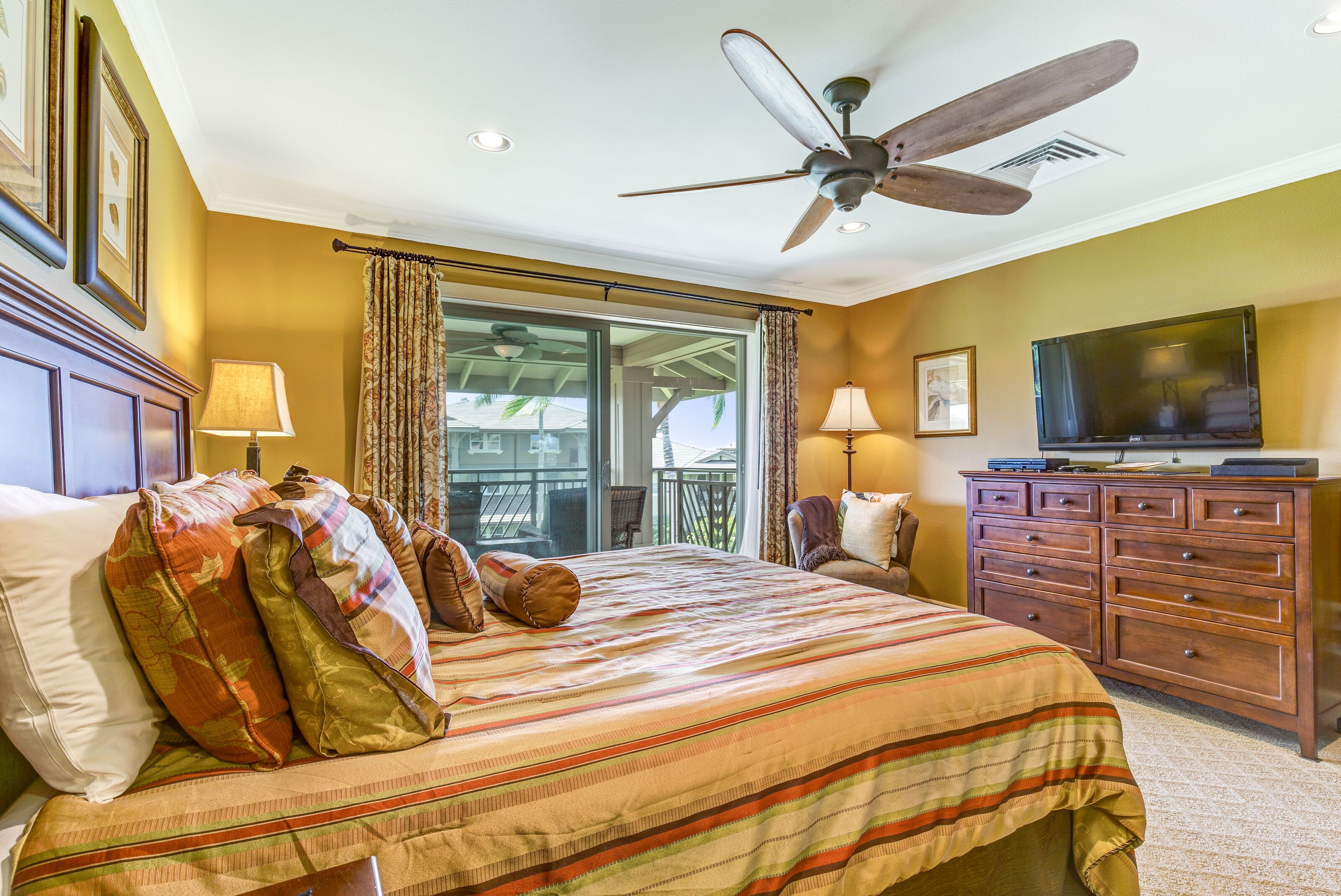 A hotel room with a bed, a television, a ceiling fan, and a view of the outdoors.