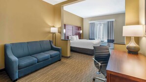 A hotel room with a blue sofa, a wooden desk, a bed with a headboard, and a large window with curtains.