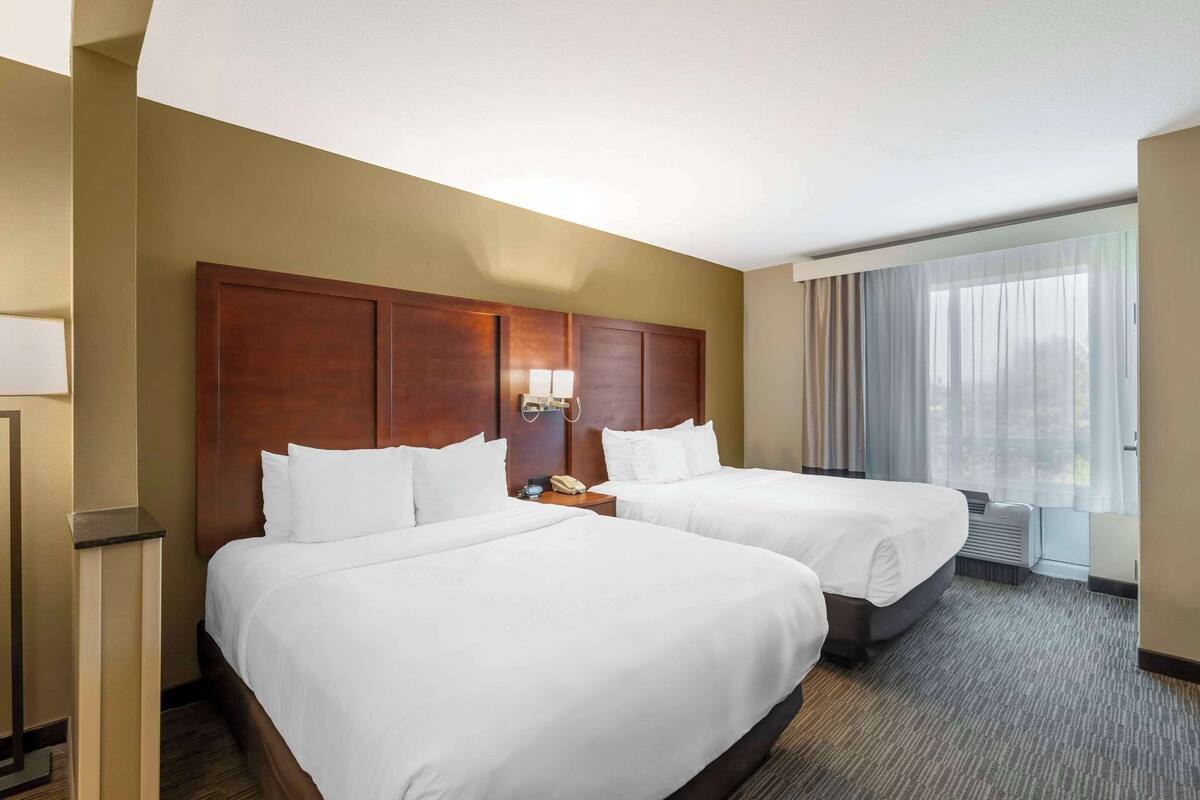 A hotel room with two beds, a wooden headboard, a bedside table with a lamp, and a large window with curtains.