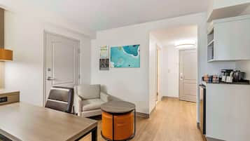 Una habitación de hotel moderna con una mesa de comedor de madera, un otoman redondo, un sofá y una zona de cocina con refrigerador y armarios.