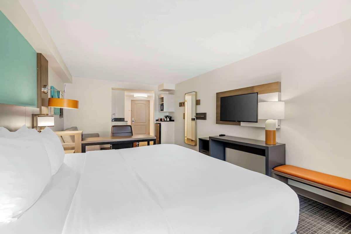 A hotel room with a large bed, a desk, a chair, a TV, and a bathroom visible in the background.