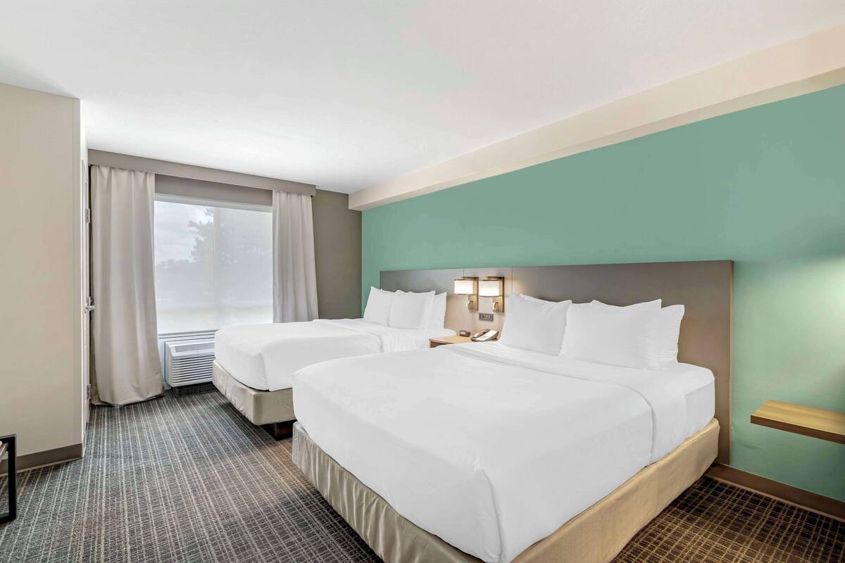 A hotel room with two beds, a large window with curtains, a bedside table with a lamp, and a wall-mounted shelf.