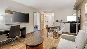 A modern hotel room with a flat-screen TV, a desk with a chair, a dining table with chairs, a sofa, and a kitchenette with a fridge and microwave. - Comfort Suites Valdosta Mall Area (Valdosta)