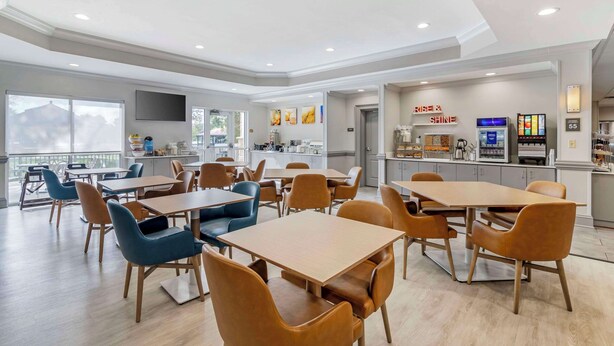 A modern dining area with tables and chairs, a kitchen area with a 'Rise & Shine' sign, and a view of the outdoors. - Comfort Suites Valdosta Mall Area (Valdosta)