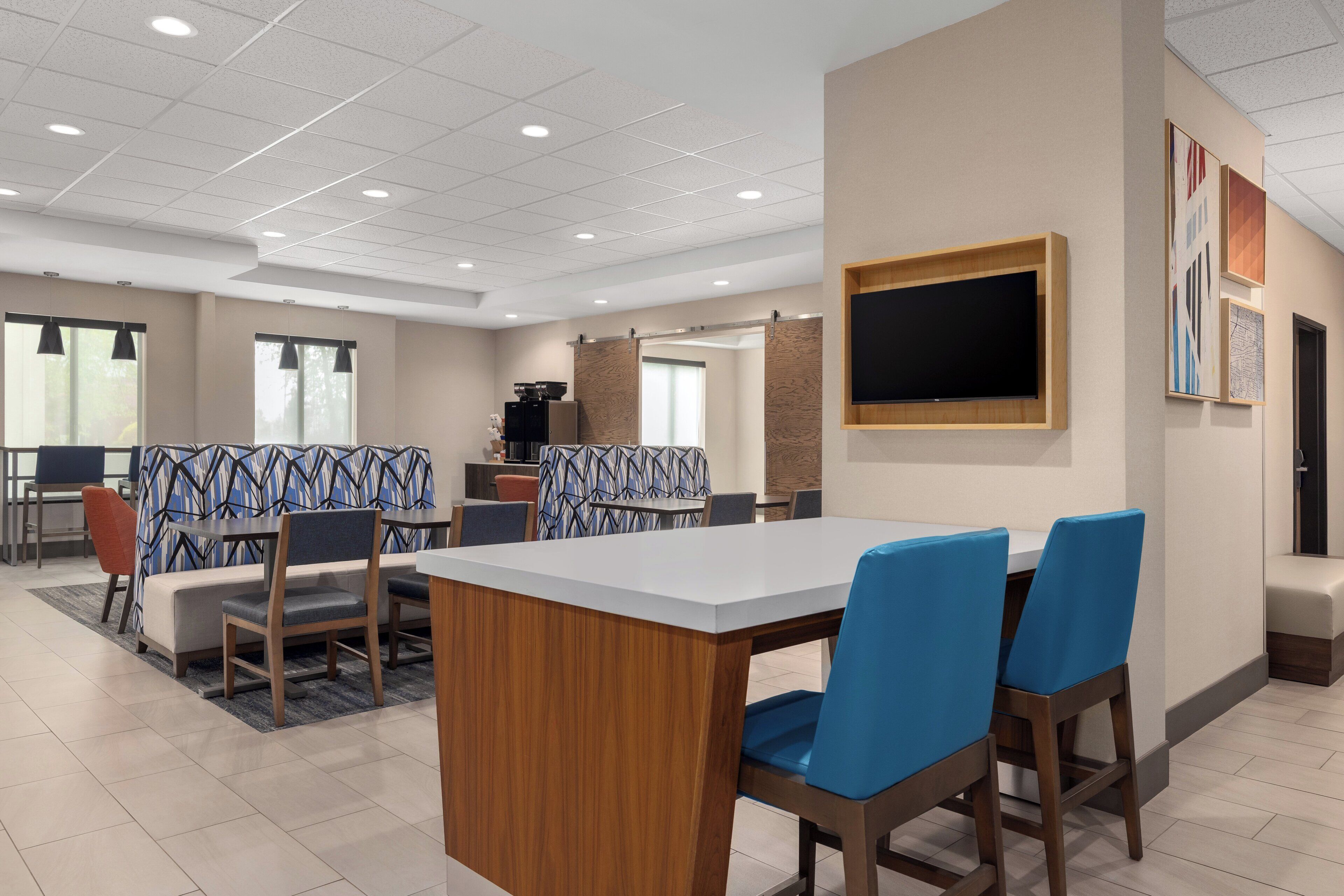 Een moderne hotel eetruimte met een grote rechthoekige tafel, blauwe zitzakstoelen, een aan de muur bevestigde televisie en een kamer met een patroon.