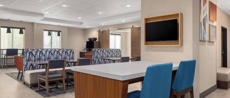 Comedor moderno de hotel con una gran mesa rectangular, sillas con acolchado azul, un televisor montado en la pared y un separador decorativo en la pared.