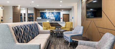 A modern hotel lobby with a sofa, armchairs, a coffee table, and a TV mounted on the wall.