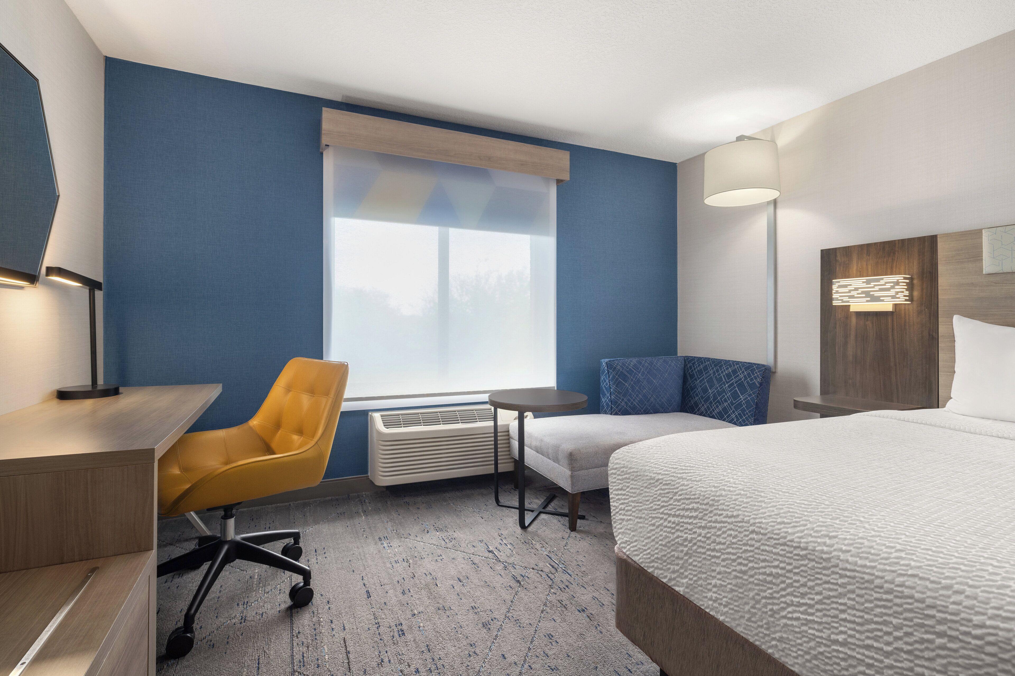 A hotel room with a bed, a desk, a chair, a small table, a heater, a window with blinds, and a wall-mounted lamp.