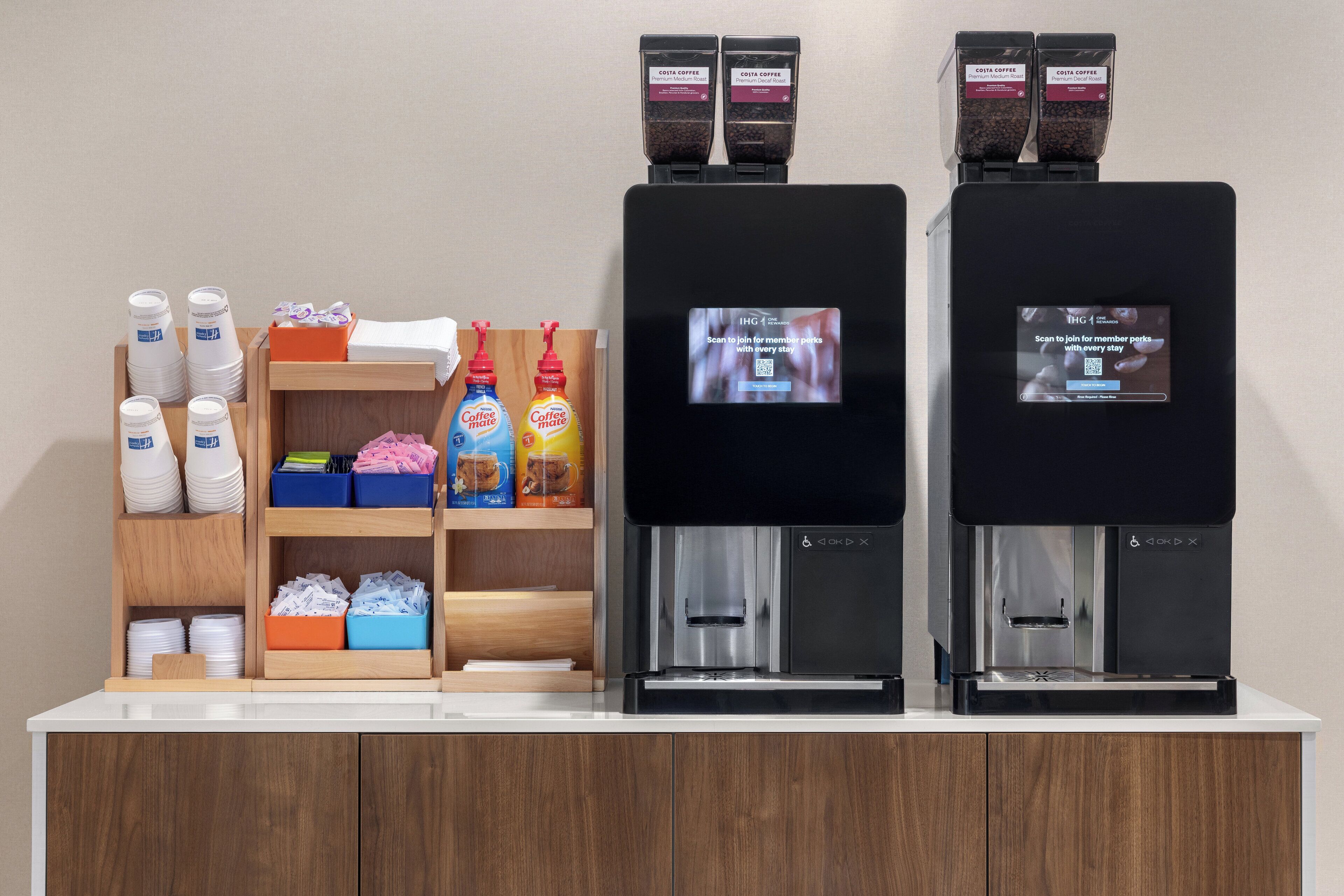 Una estación de café con dos dispensadores, una repisa con sobres de café y una exhibición de vasos y platos desechables.