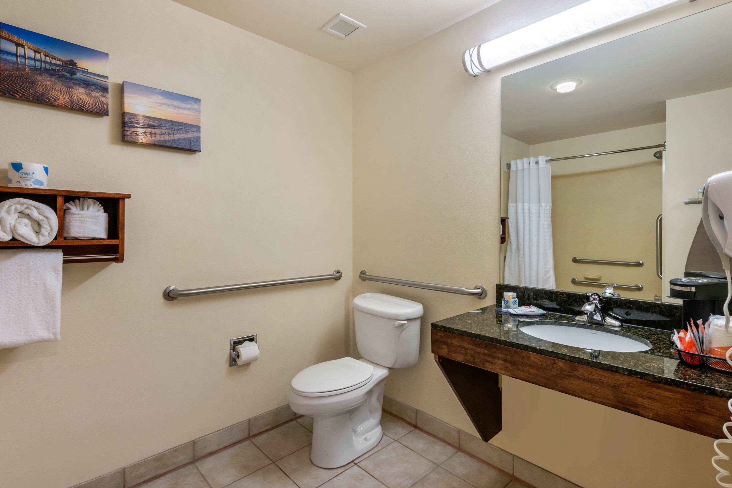 Baño con inodoro, lavabo y ducha. Cuenta con barras de apoyo, toallero y espejo.