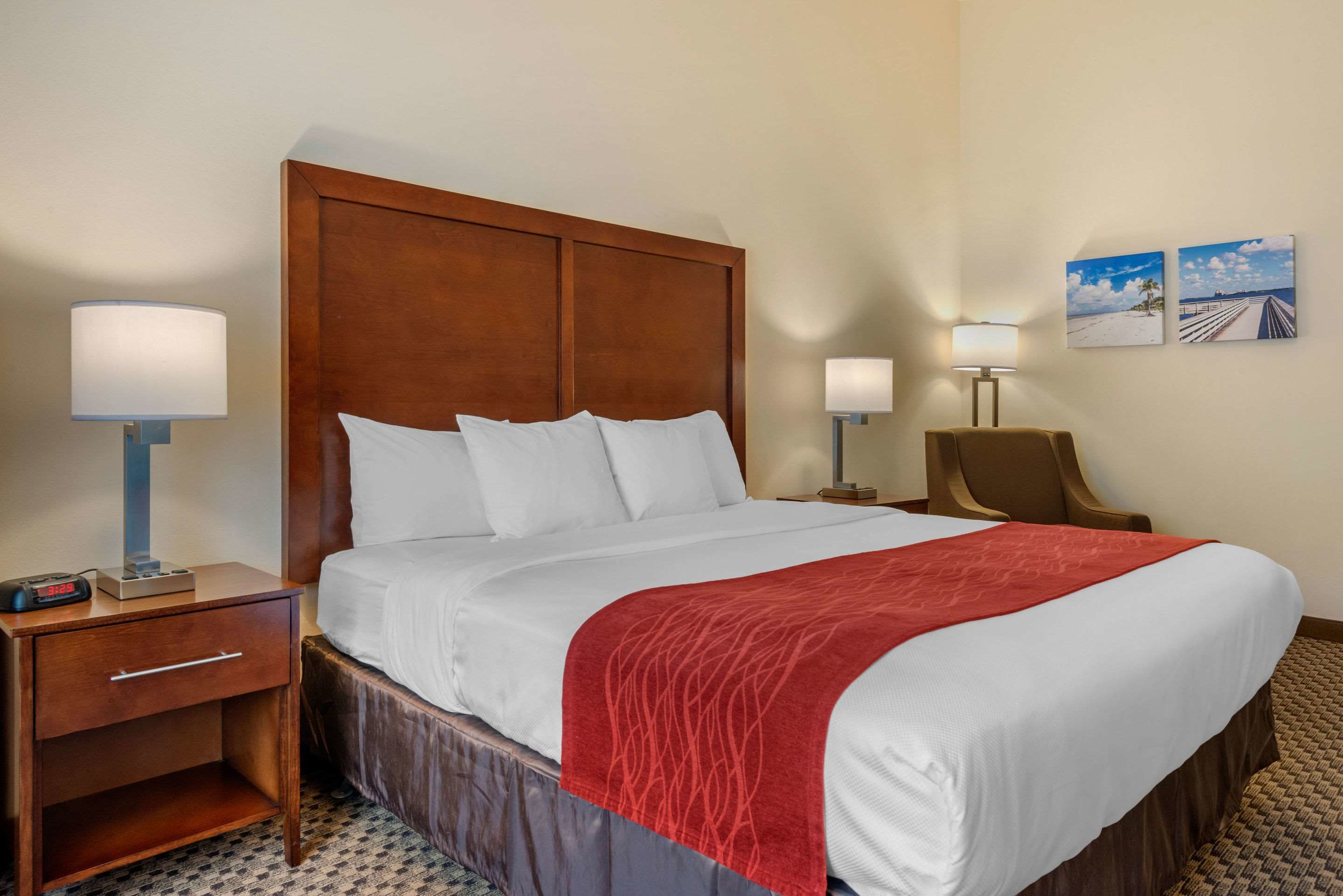 A hotel room with a large bed, wooden headboard, two bedside lamps, a nightstand with a lamp, and two framed pictures on the wall.
