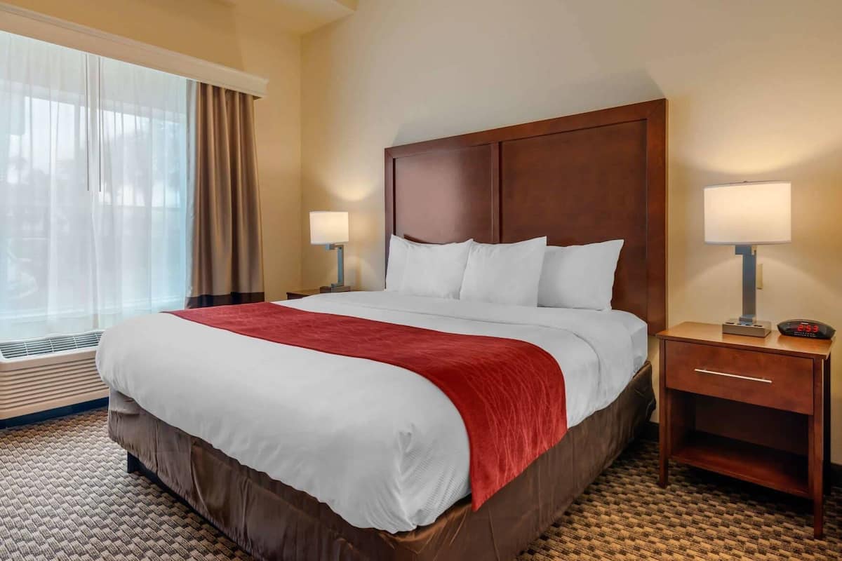 A hotel room with a large bed, two bedside tables, a nightstand, a window with curtains, and an air conditioning unit.