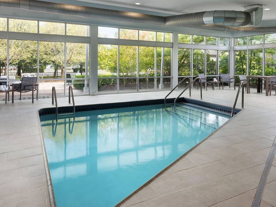An indoor swimming pool with clear glass walls, surrounded by seating areas and a view of trees.