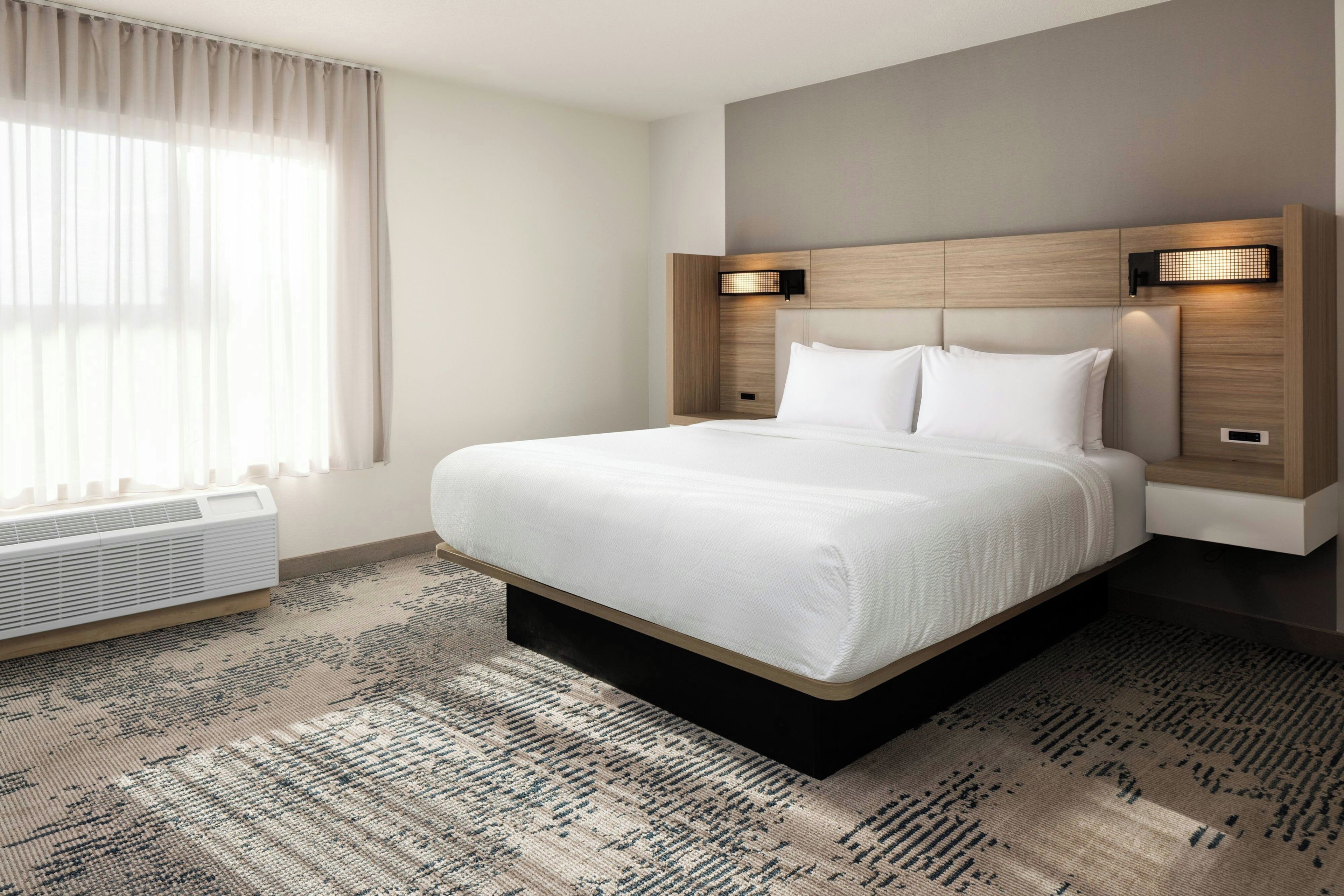 A hotel room with a large bed, a bedside table, a wall-mounted light, and a window with curtains.