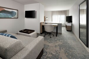Habitación de hotel con sofá, escritorio con silla, televisor de pantalla plana montado en la pared y un espejo grande que refleja la habitación.