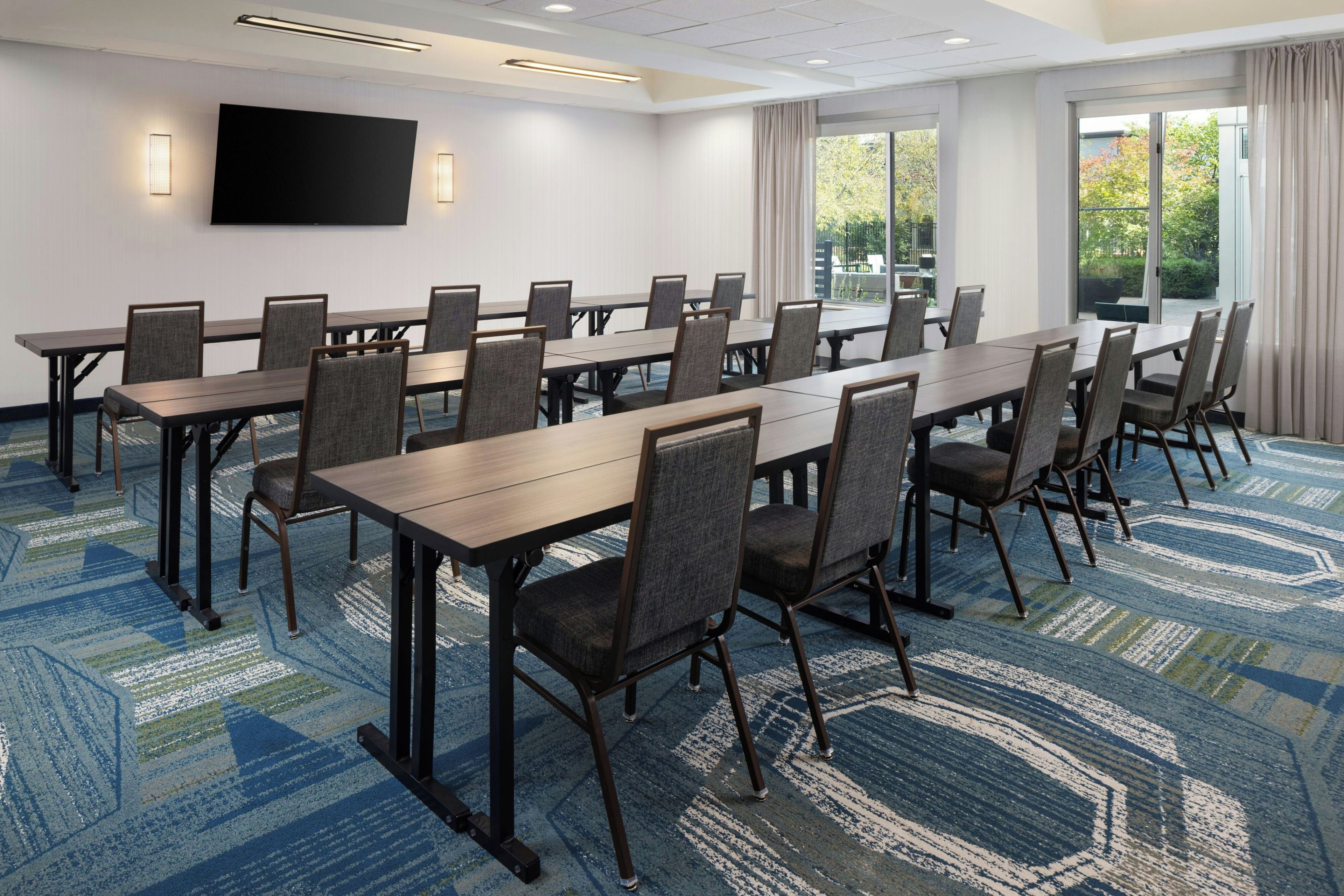 A conference room with multiple tables and chairs, a flat-screen TV mounted on the wall, and large windows with curtains.