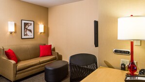 A hotel room with a beige sofa, a red pillow, a black chair, a wooden table, a red lamp, and a wall clock. - SpringHill Suites by Marriott Appleton (Appleton)