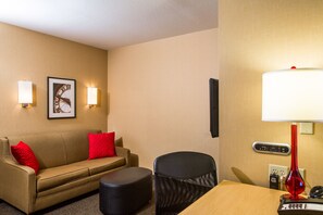 A hotel room with a beige sofa, a red pillow, a black chair, a wooden table, a red lamp, and a wall clock. - Cambria Hotel Appleton (Appleton)
