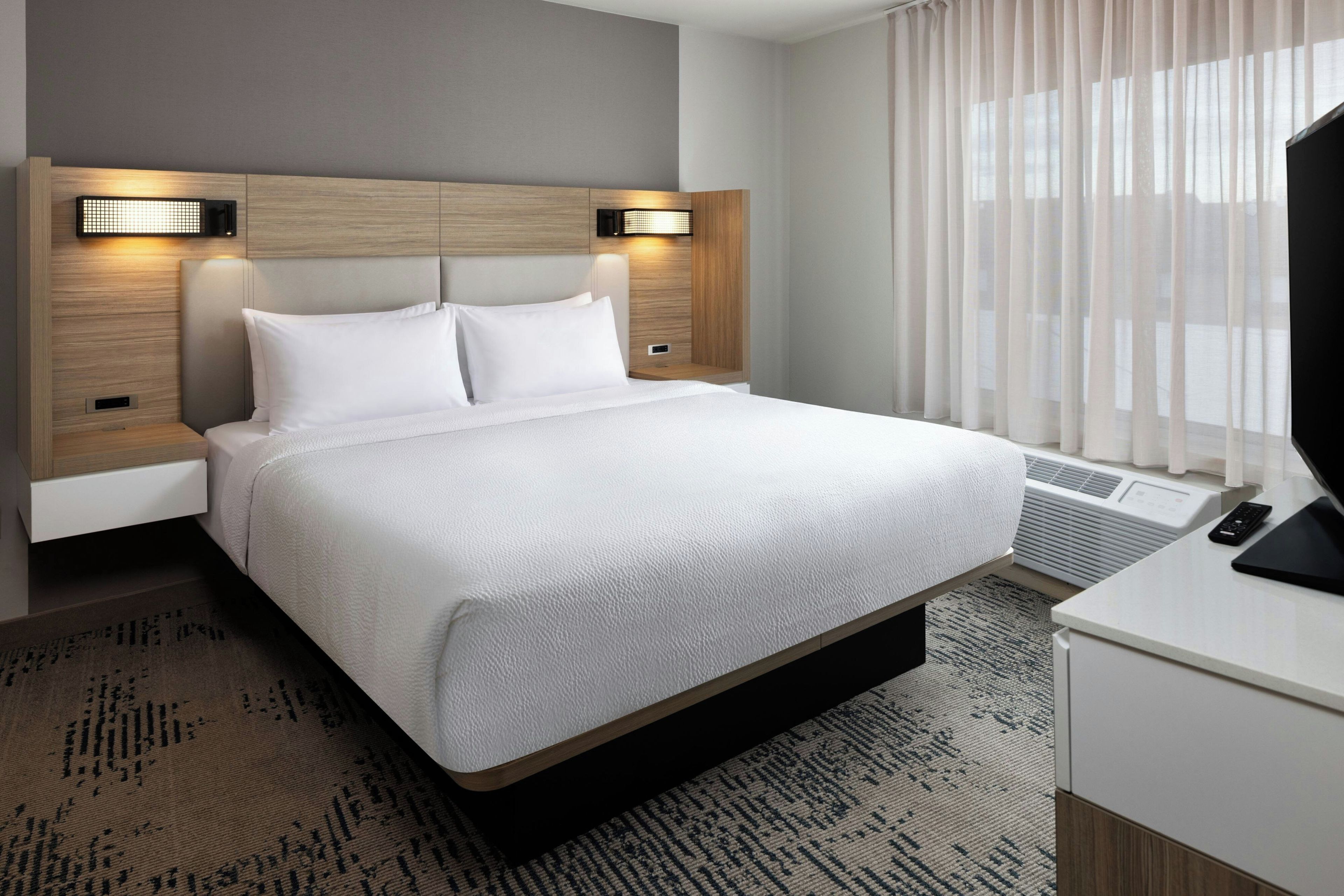 Habitación de hotel con una cama grande, una mesita de noche, un televisor y una ventana con cortinas.