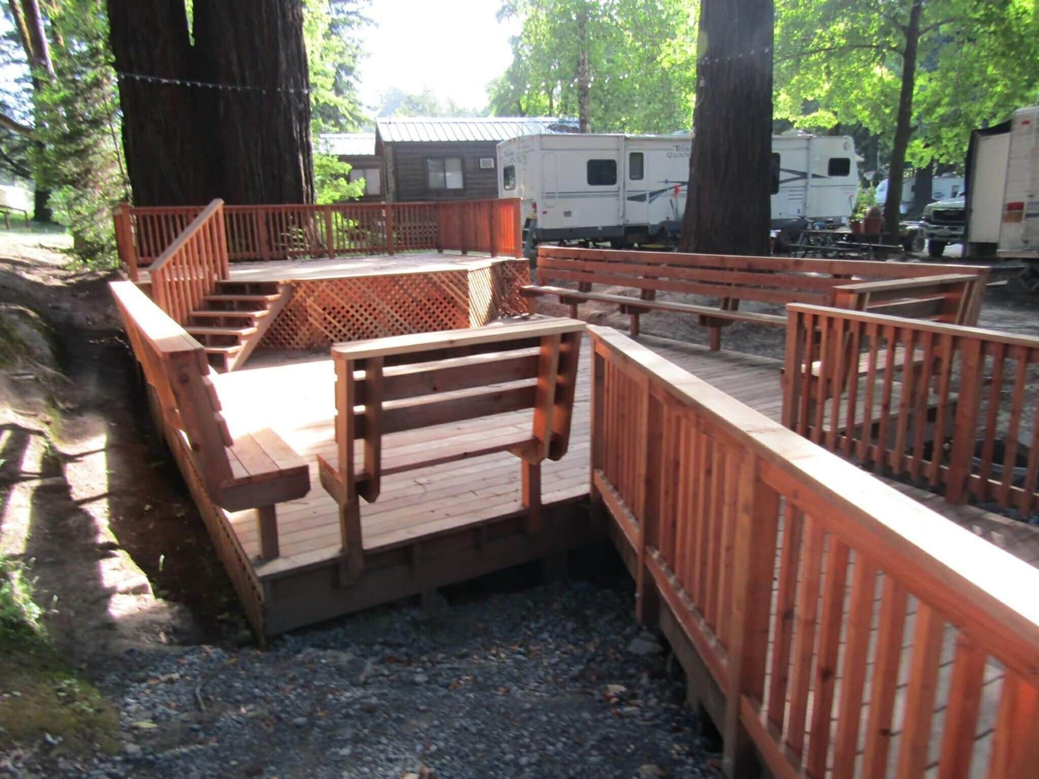 A wooden deck with benches and picnic tables, surrounded by trees and RVs.