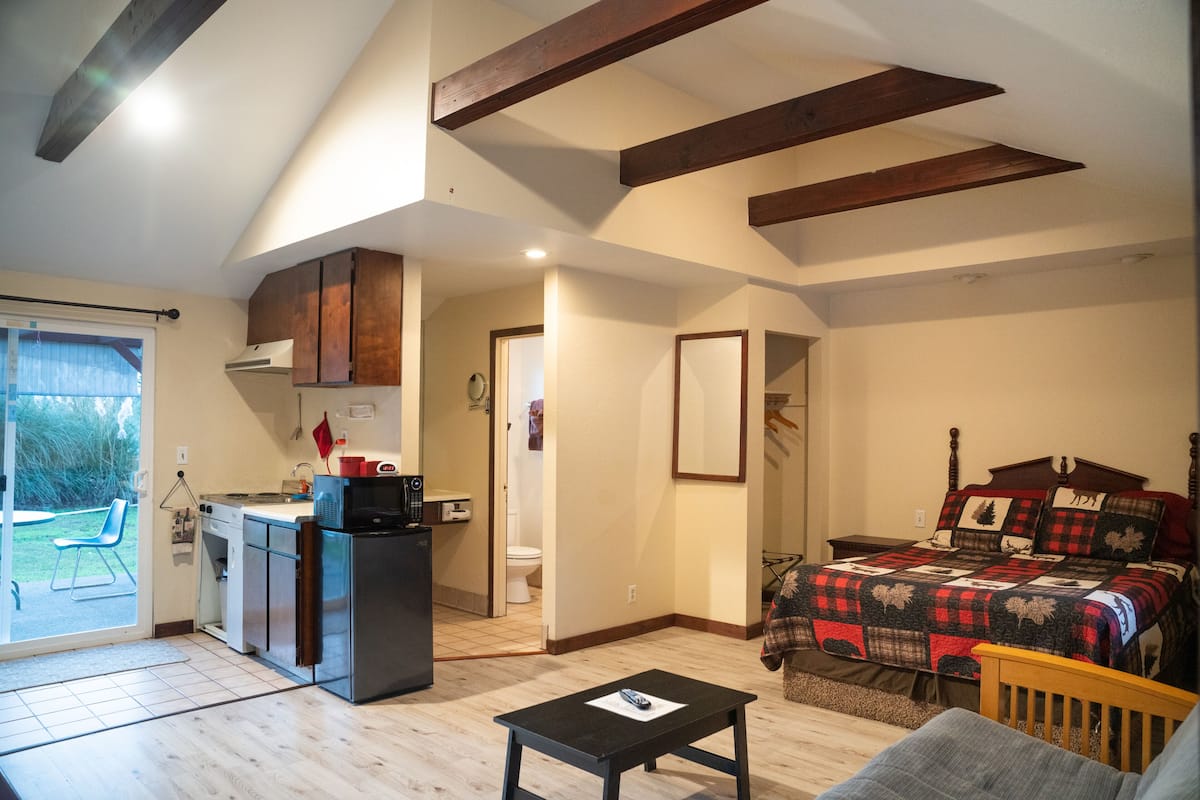 A compact room with a kitchenette, a bed with a quilt, a small table, and a view of the outside.
