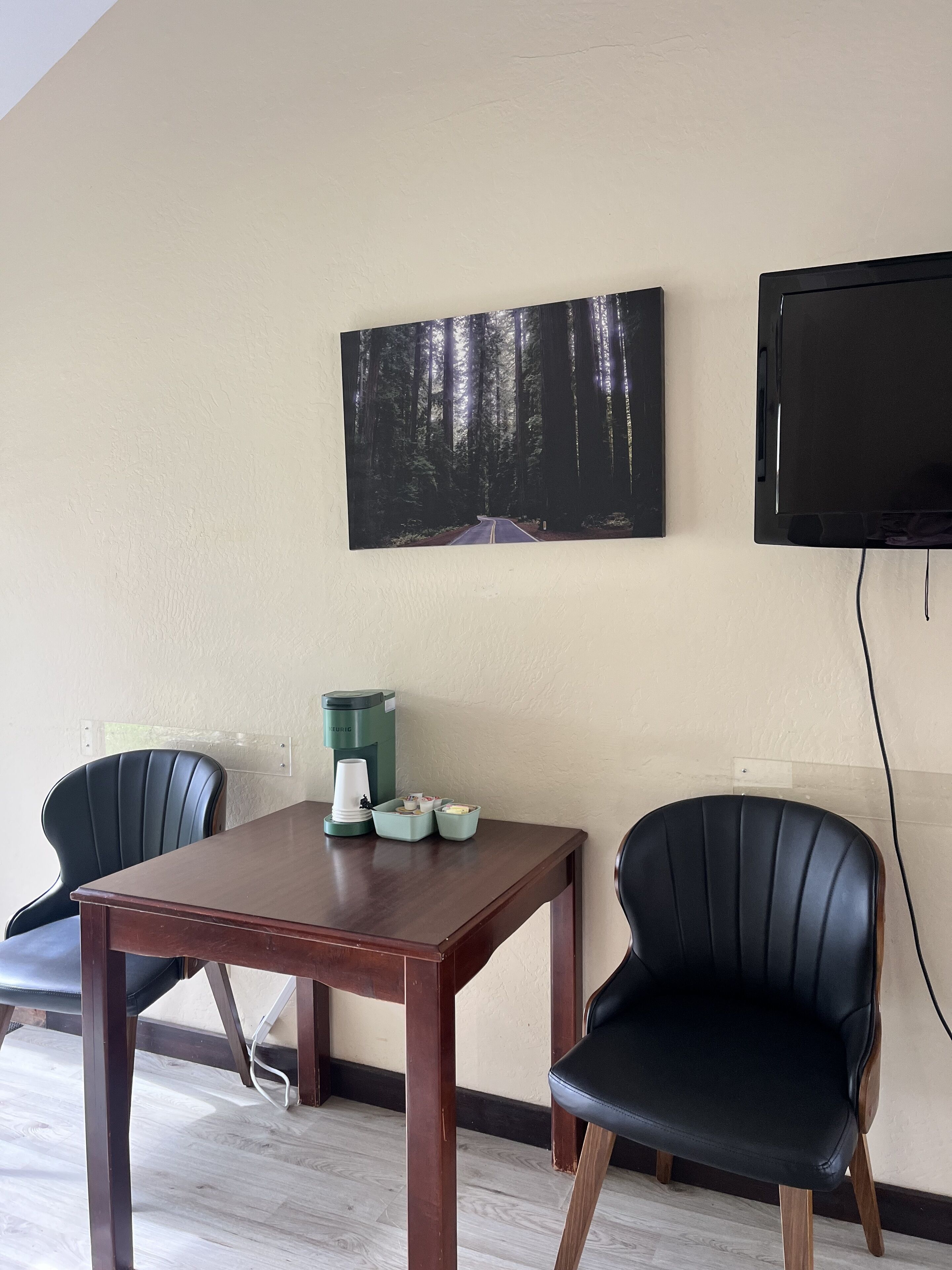 Une petite pièce avec une table en bois, deux chaises noires, une machine à café et un tableau représentant une forêt accroché au mur.