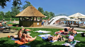 Een park met een tuinhuisje met rieten dak, picknicktafels en mensen die op dekens zonnen.