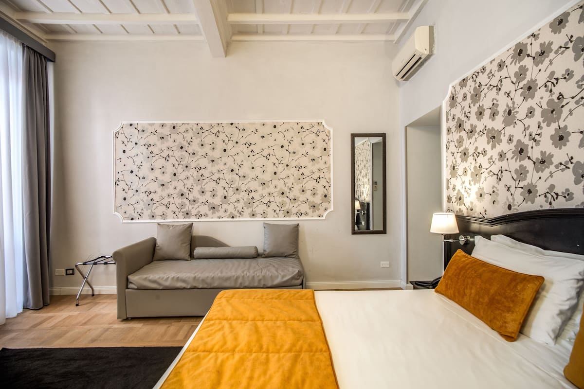 A hotel room with a bed, a sofa, a large floral wallpaper, a mirror, and a window with curtains.