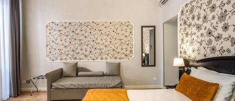 Habitación de hotel con cama, sofá, papel tapiz con estampado floral, espejo y ventana con cortinas.