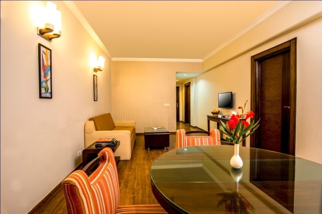 A hotel room with a round glass table, striped chairs, a sofa, a TV, and a vase with flowers.