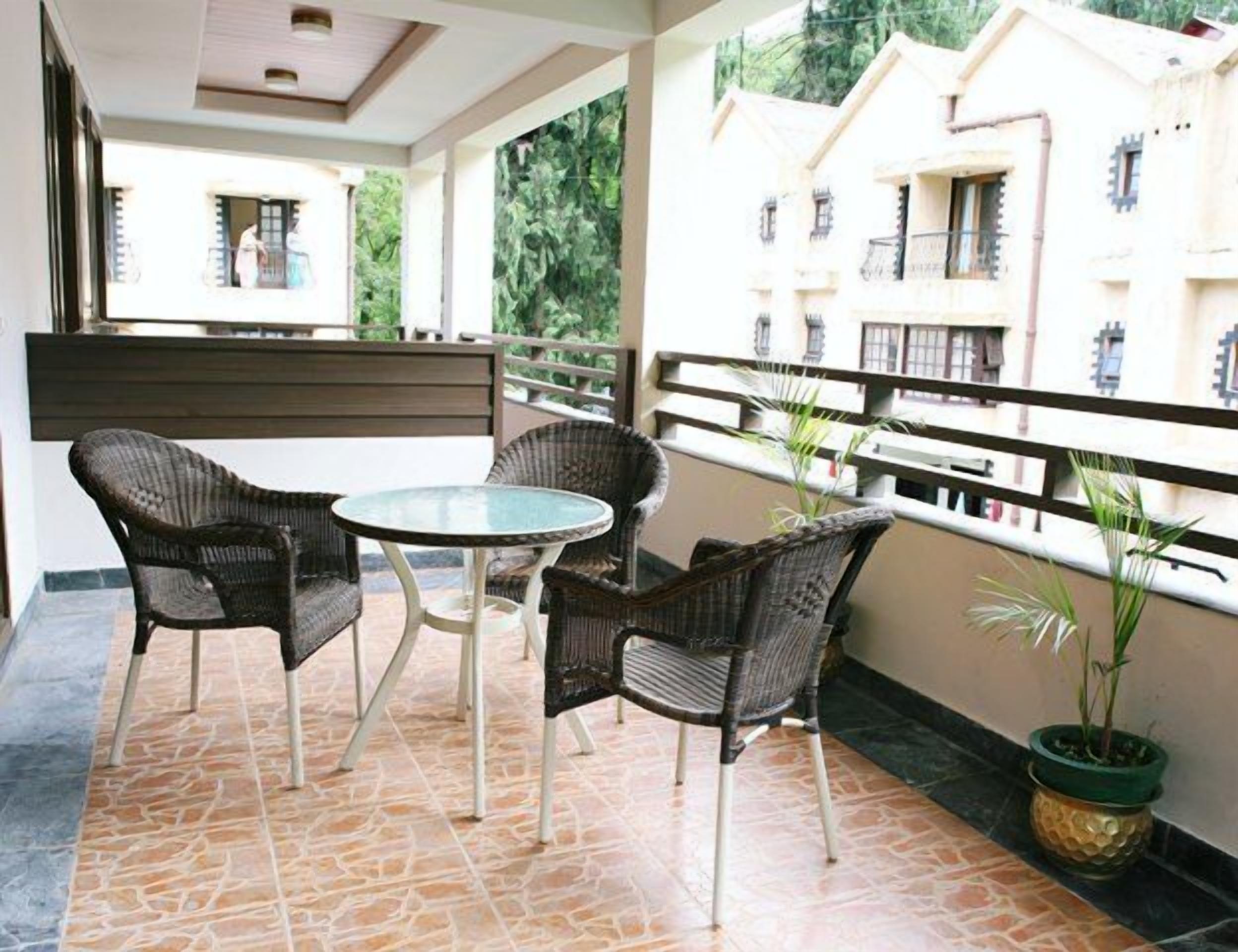 A balcony with wicker furniture and a glass table, overlooking a residential area with buildings and greenery.