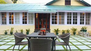 A patio with a table and four wicker chairs, a house with a green roof, and potted plants.