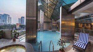 A modern indoor swimming pool with a view of the cityscape, a hot tub, and a lounge area with a striped towel. - The Regenza by Tunga (Navi Mumbai)