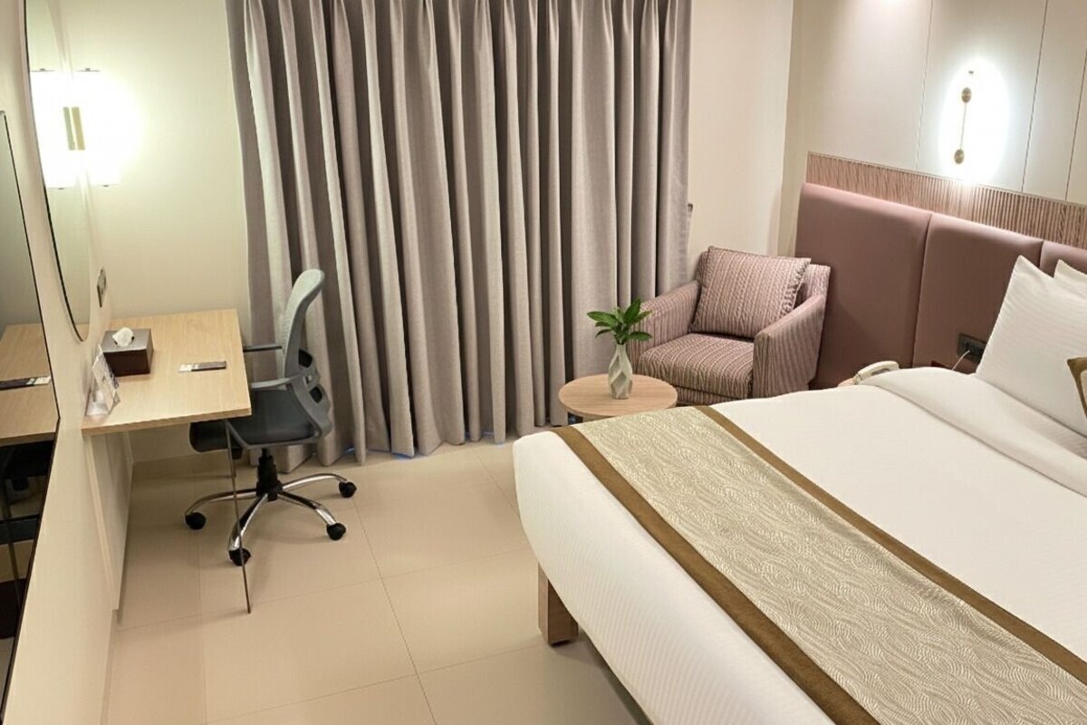 A hotel room with a bed, a desk, a chair, a small table with a plant, and curtains.