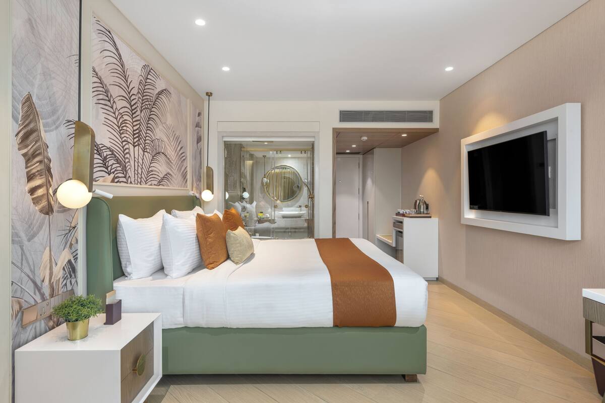 A modern hotel room with a large bed, a TV mounted on the wall, and a bathroom visible through a glass door.