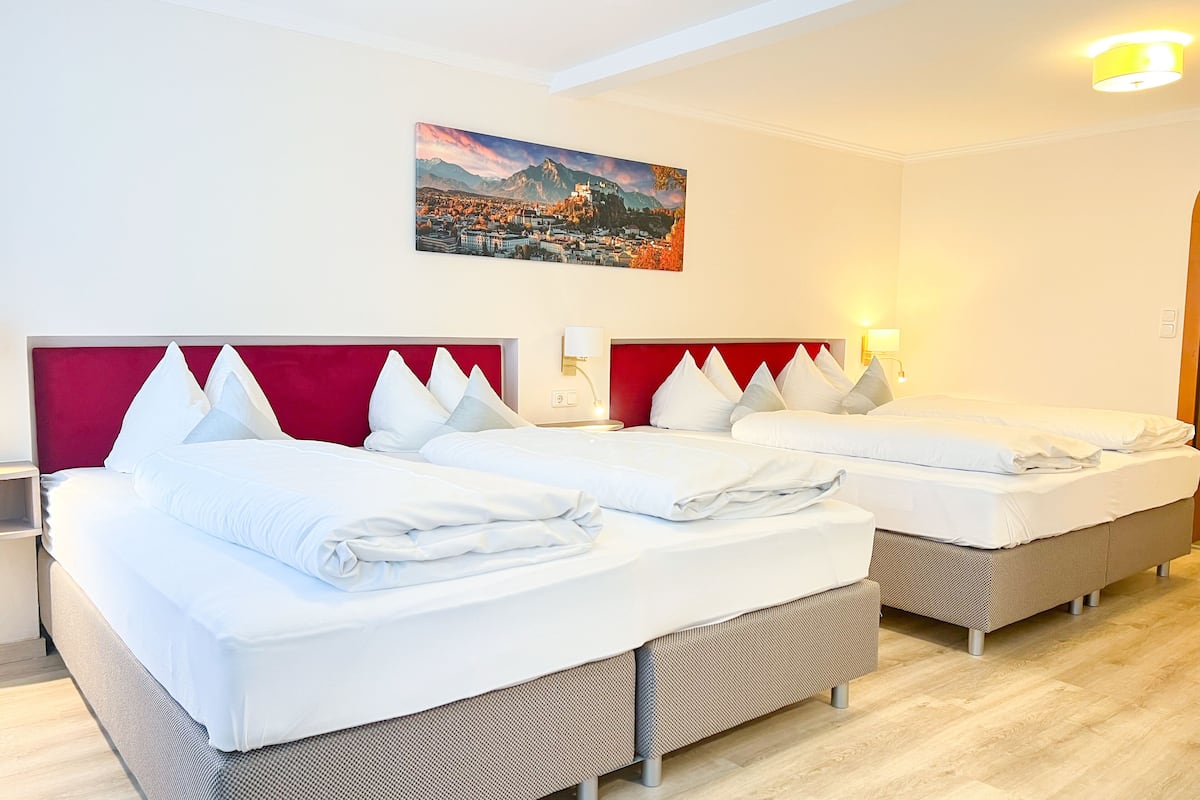 Ein Hotelzimmer mit zwei Betten, einem roten Kopfteil, weißer Bettwäsche, einem Gemälde einer Bergstadt und einem Nachttisch mit Lampe.