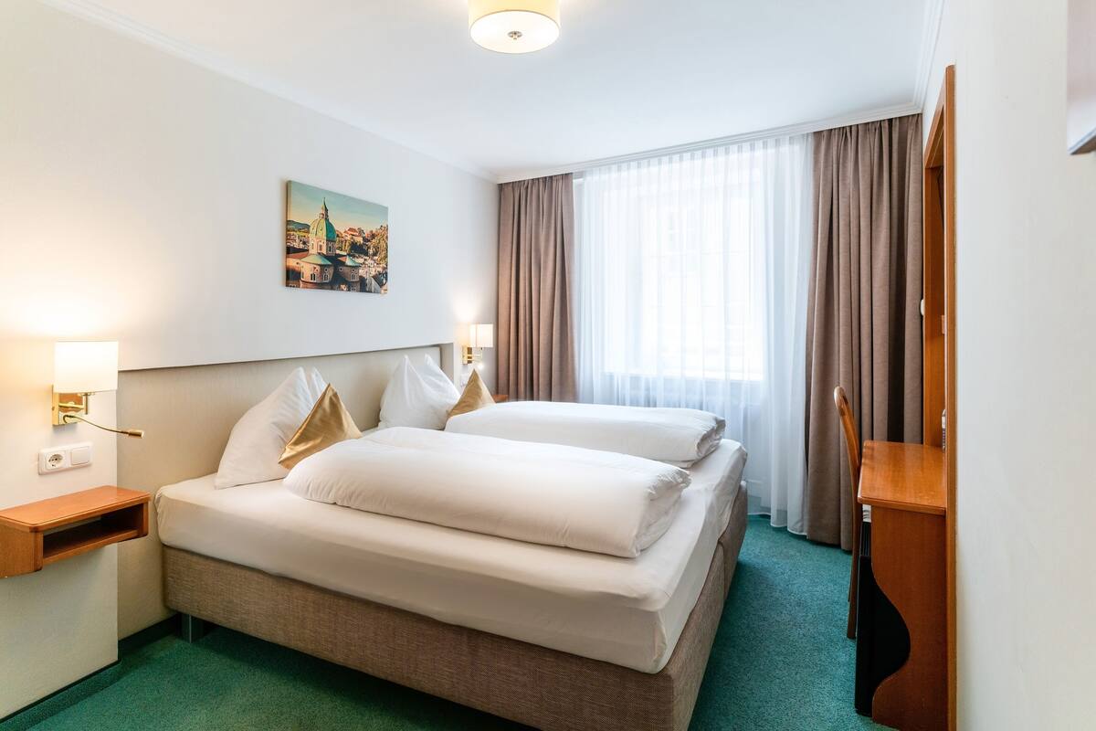 Ein Hotelzimmer mit Doppelbett, Nachttisch, Lampe, einem Bild an der Wand und einem Fenster mit Vorhängen.