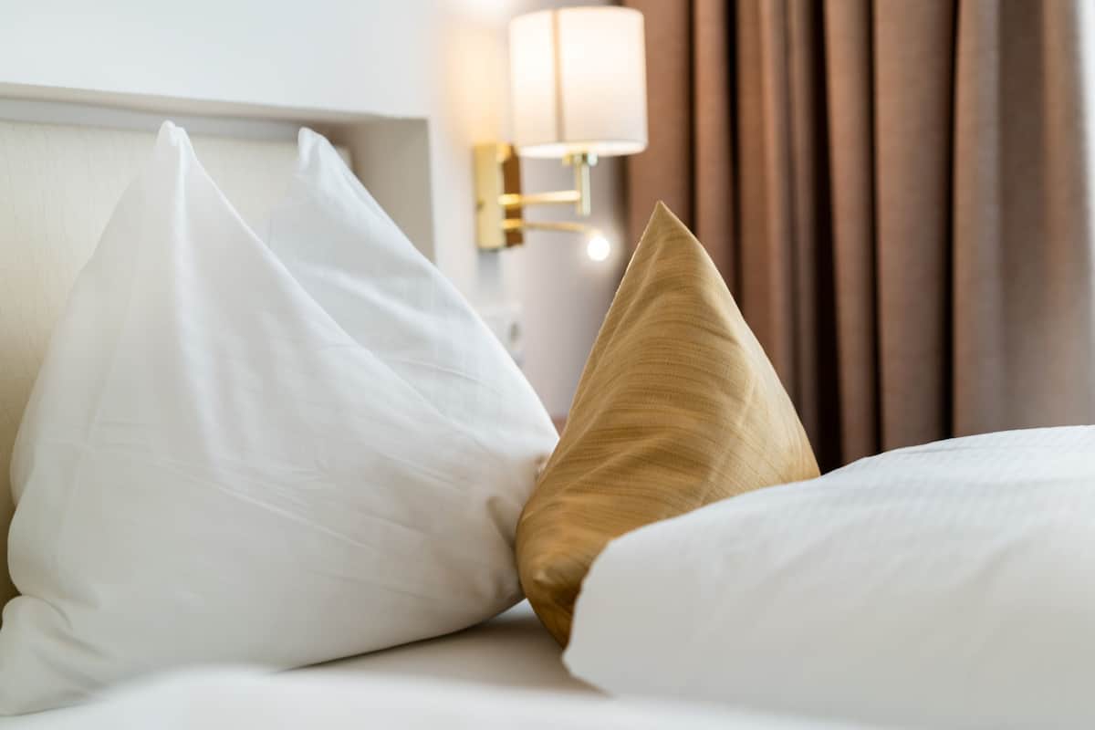 Ein ordentlich bezogenes Bett mit weißen und goldenen Kissen, einer Nachttischlampe und einem Vorhang im Hintergrund.