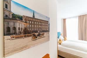 Une chambre d’hôtel avec un grand tableau historique représentant une scène de rue, un lit avec du linge de maison blanc et une fenêtre avec des rideaux.