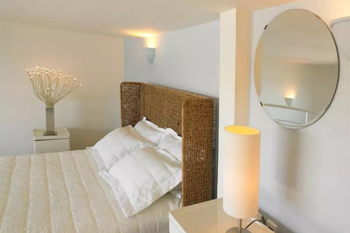 A neatly made bed with white linens, a woven headboard, a bedside table with a lamp, and a round mirror on the wall.