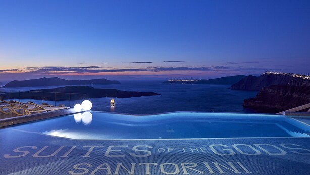 Una piscina infinita con el texto "Suites of the Gods Santorini", con vistas a un paisaje costero.
