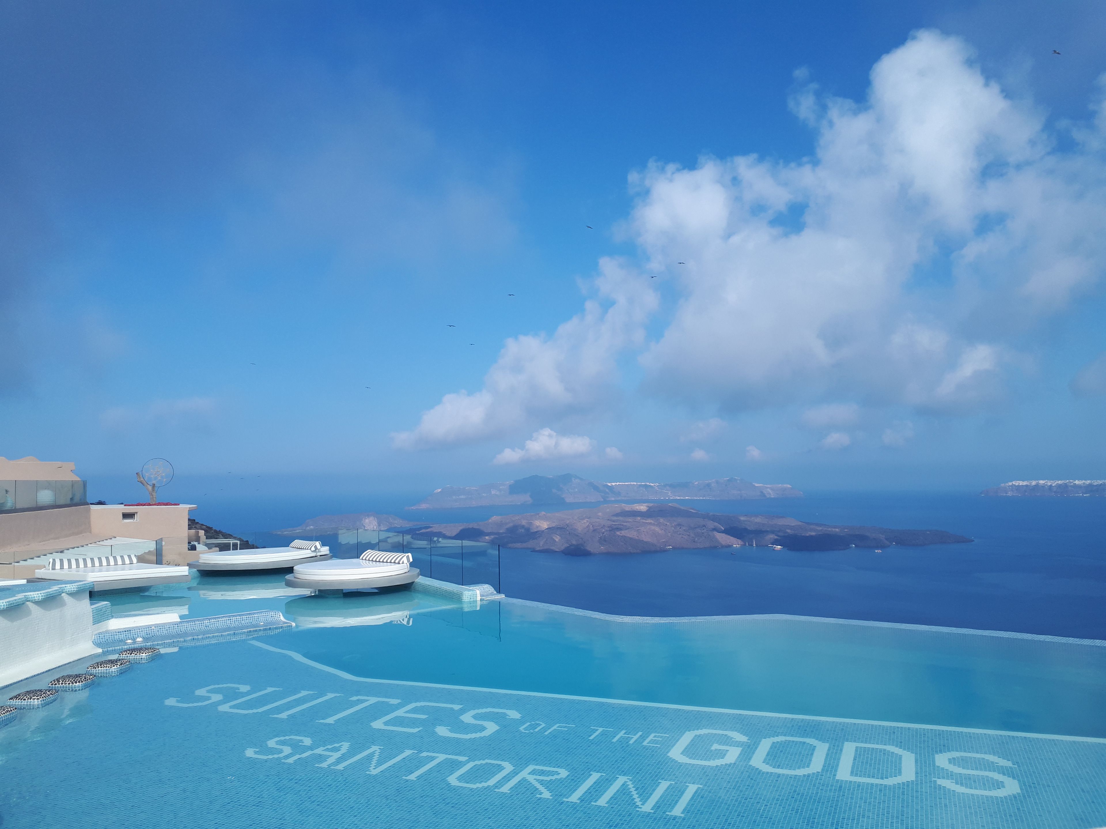 Une magnifique piscine à débordement d’un bleu profond, avec l’inscription « Suites of the Gods Santorini » et une vue sur la mer et les îles en arrière-plan.